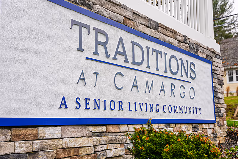 Stone entrance sign reading "Traditions at Camargo — A Senior Living Community" with landscaping in front.