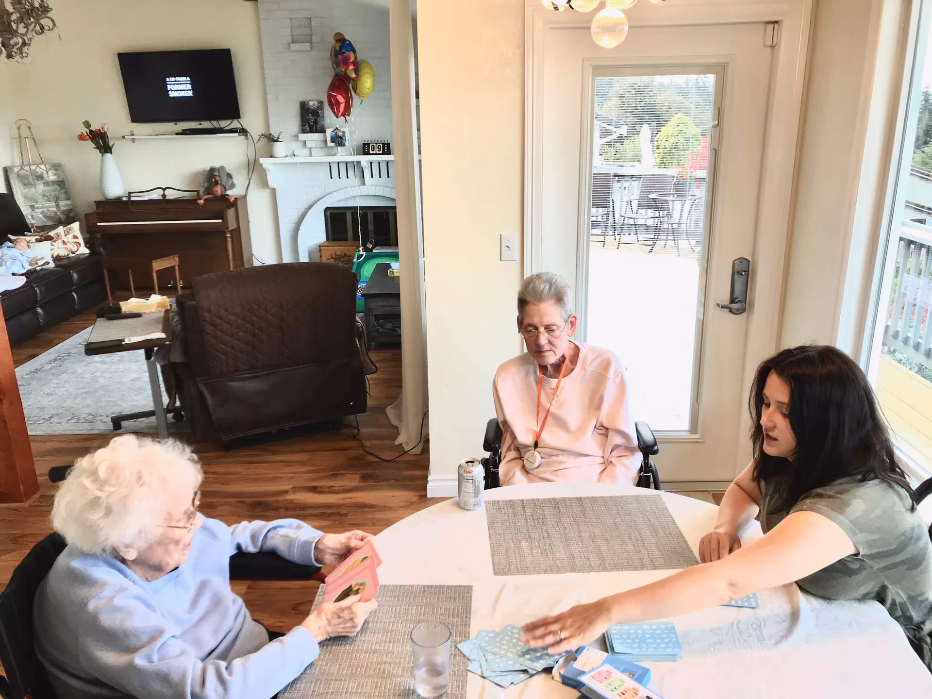 Three people sit around a round table playing cards in a bright common room with a door to an outdoor deck and furniture including a piano and TV in the background.
