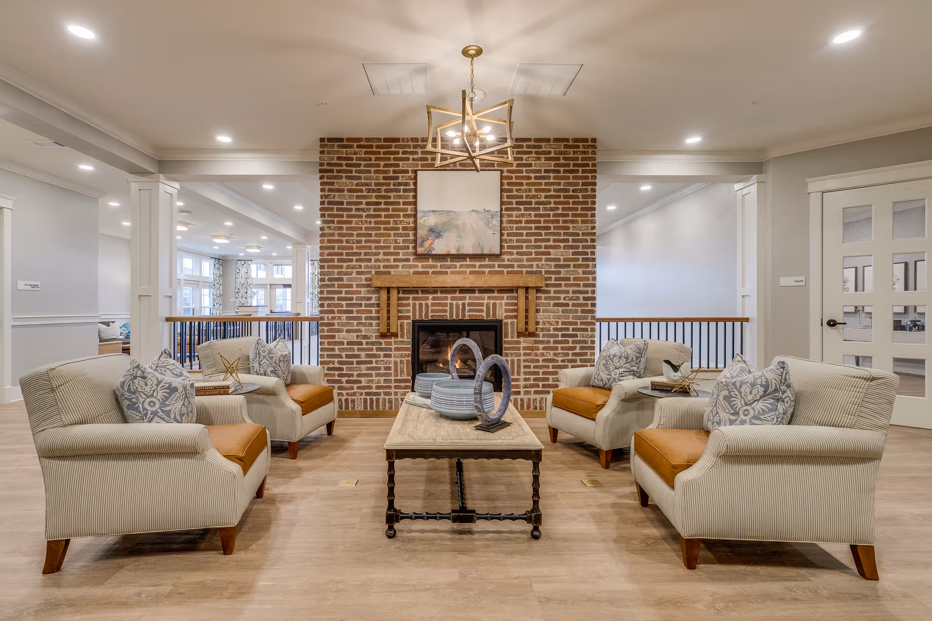 A cozy living room area with four beige armchairs featuring orange cushions and patterned pillows arranged around a wooden coffee table. The coffee table holds decorative items including plates and circular sculptures. A brick fireplace with a wooden mantle and a framed painting above it is centered on the back wall. The room has light wood flooring, white walls, and recessed ceiling lights, with a modern chandelier hanging above the coffee table.