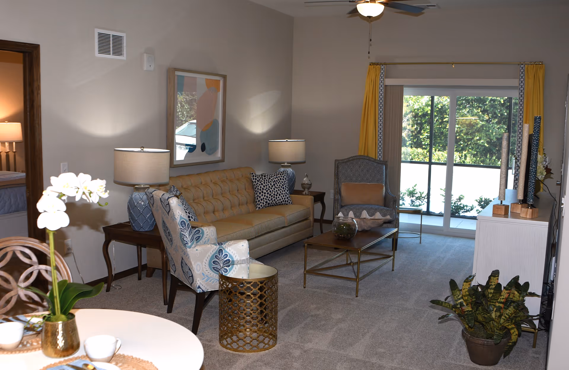 Bright living room in a retirement community with a sofa, armchairs, coffee table, lamps, sliding glass doors and a dining table in the foreground.