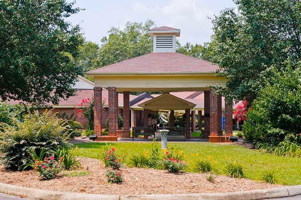 Front entrance of a senior living facility with a covered porte-cochère supported by brick columns, landscaped circular driveway and a small decorative fountain.