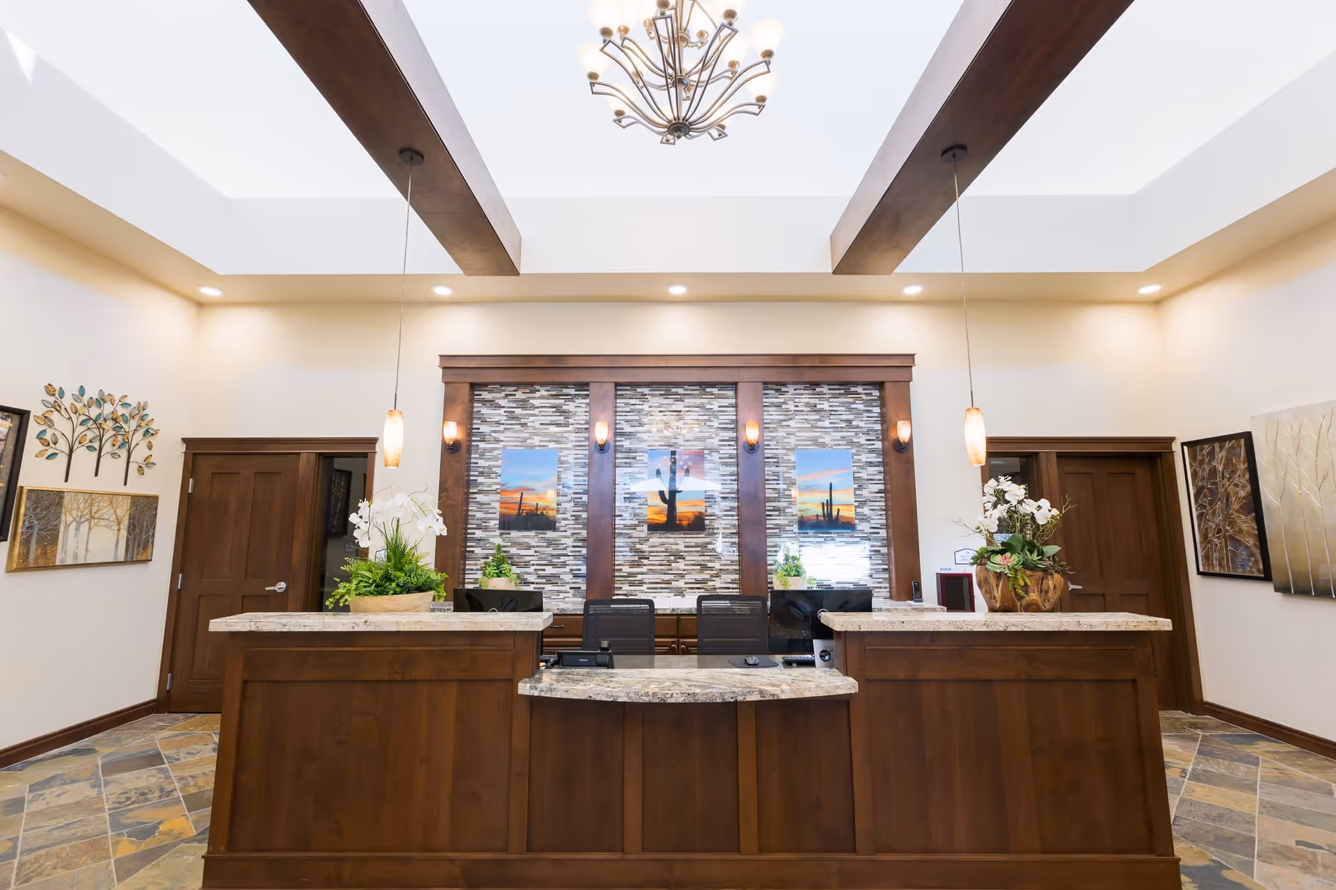 Reception area with a wooden front desk featuring granite countertops, two pendant lights hanging from the ceiling, and decorative plants on the desk. Behind the desk is a wall with a mosaic tile design and three framed pictures of desert landscapes with cacti. The room has a high ceiling with wooden beams and a chandelier, and there are two wooden doors on either side of the desk. The walls are decorated with framed artwork and a metal tree sculpture.