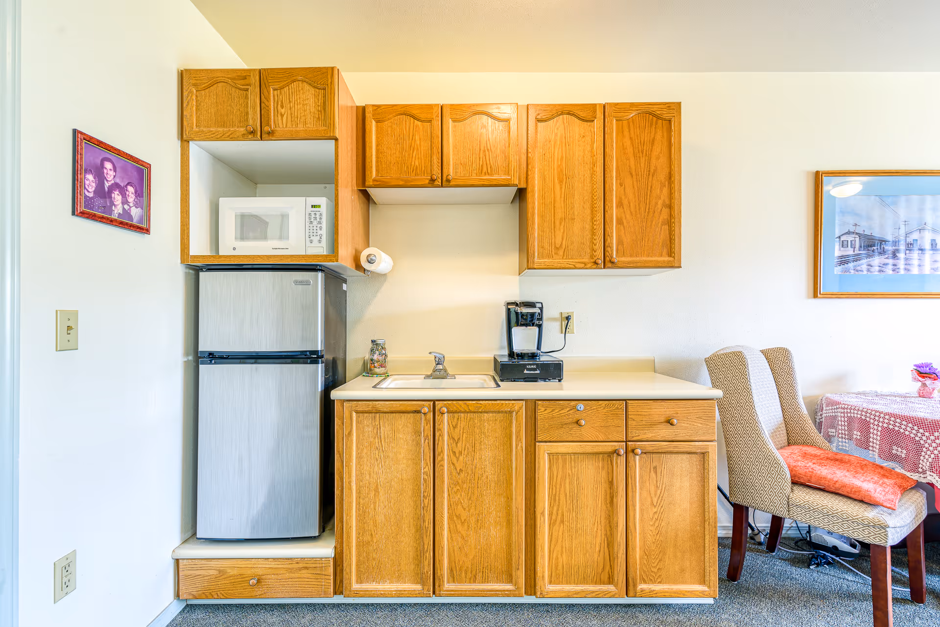 Small kitchenette with wooden cabinets, compact refrigerator, microwave, sink, and coffee maker beside a dining chair and table.