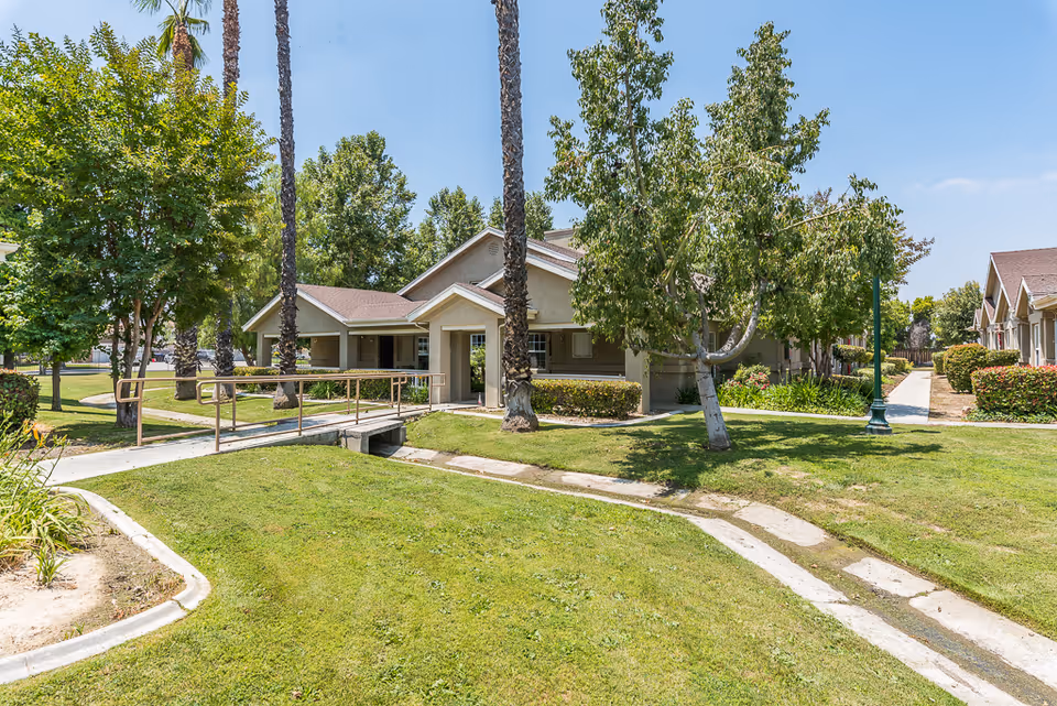 Front exterior of a single-story cottage with a wheelchair ramp, lawn and trees on a sunny day.