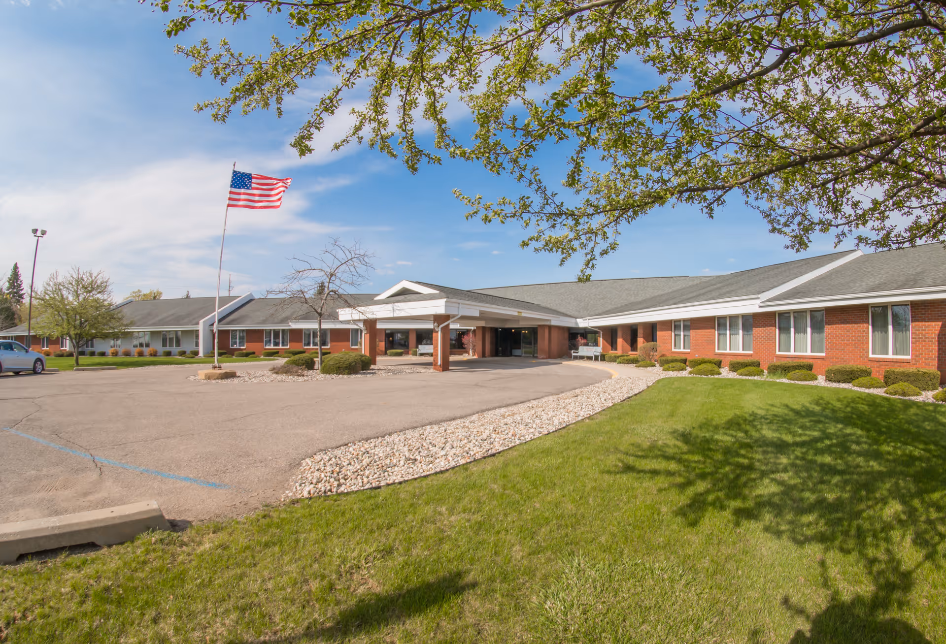 Exterior view of a single-story senior living facility building with a covered entrance, an American flag on a flagpole, a parking area, and green lawn with trees under a blue sky.