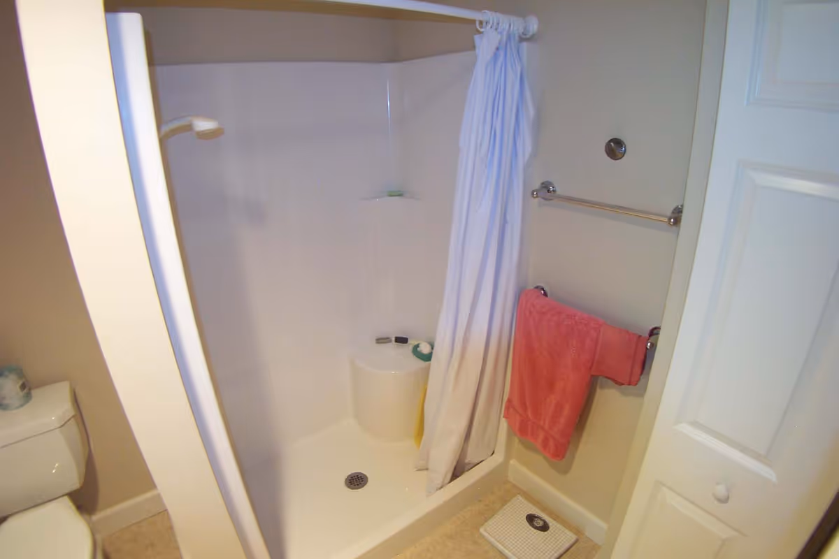 Corner shower stall with a white curtain in a small bathroom, a toilet to the left, a red towel on a wall rack and a scale on the floor.
