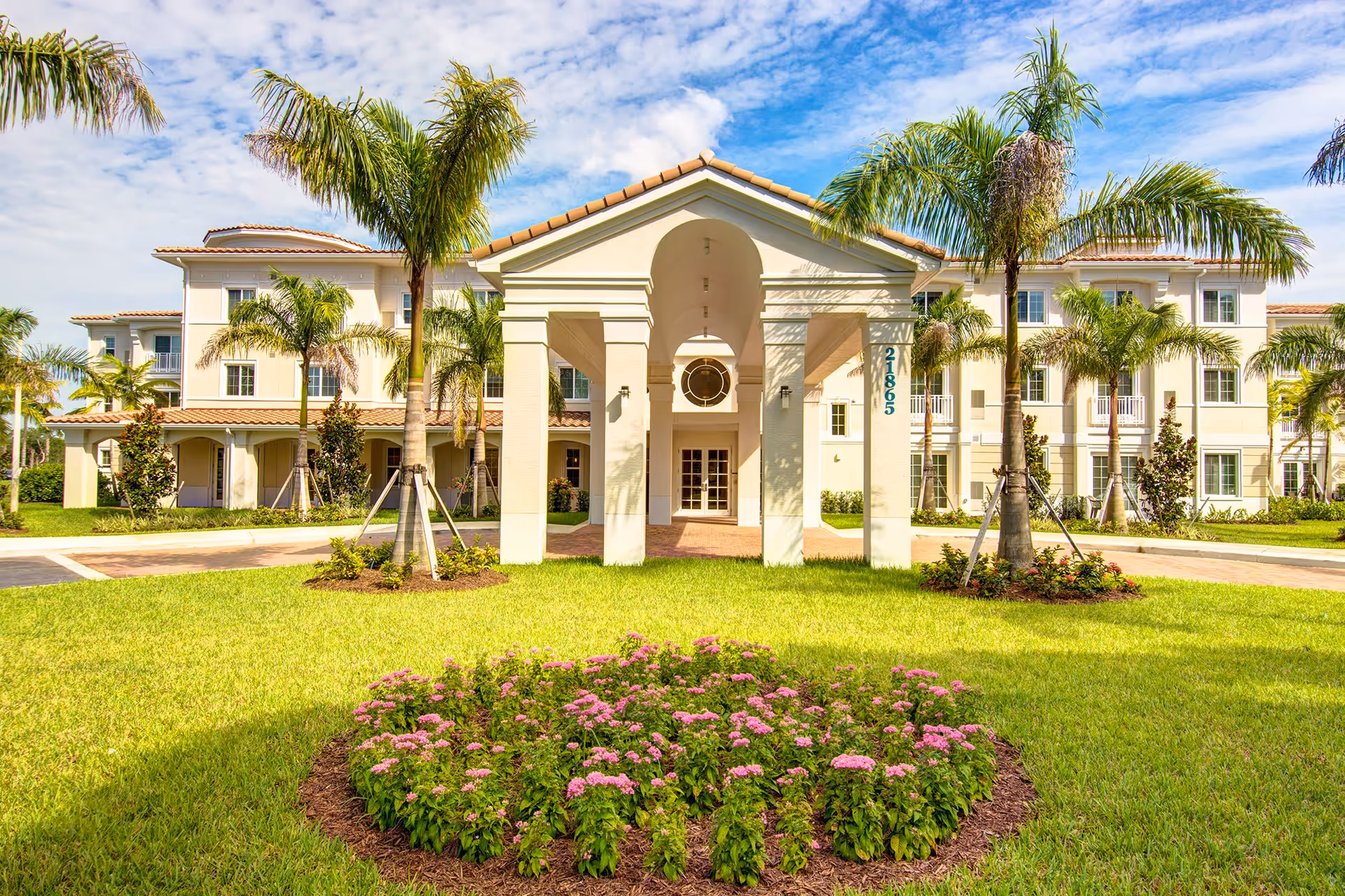 Front exterior view of The Meridian at Boca Raton building with a covered entrance, palm trees, green lawn, and a flower bed with pink flowers under a partly cloudy blue sky.
