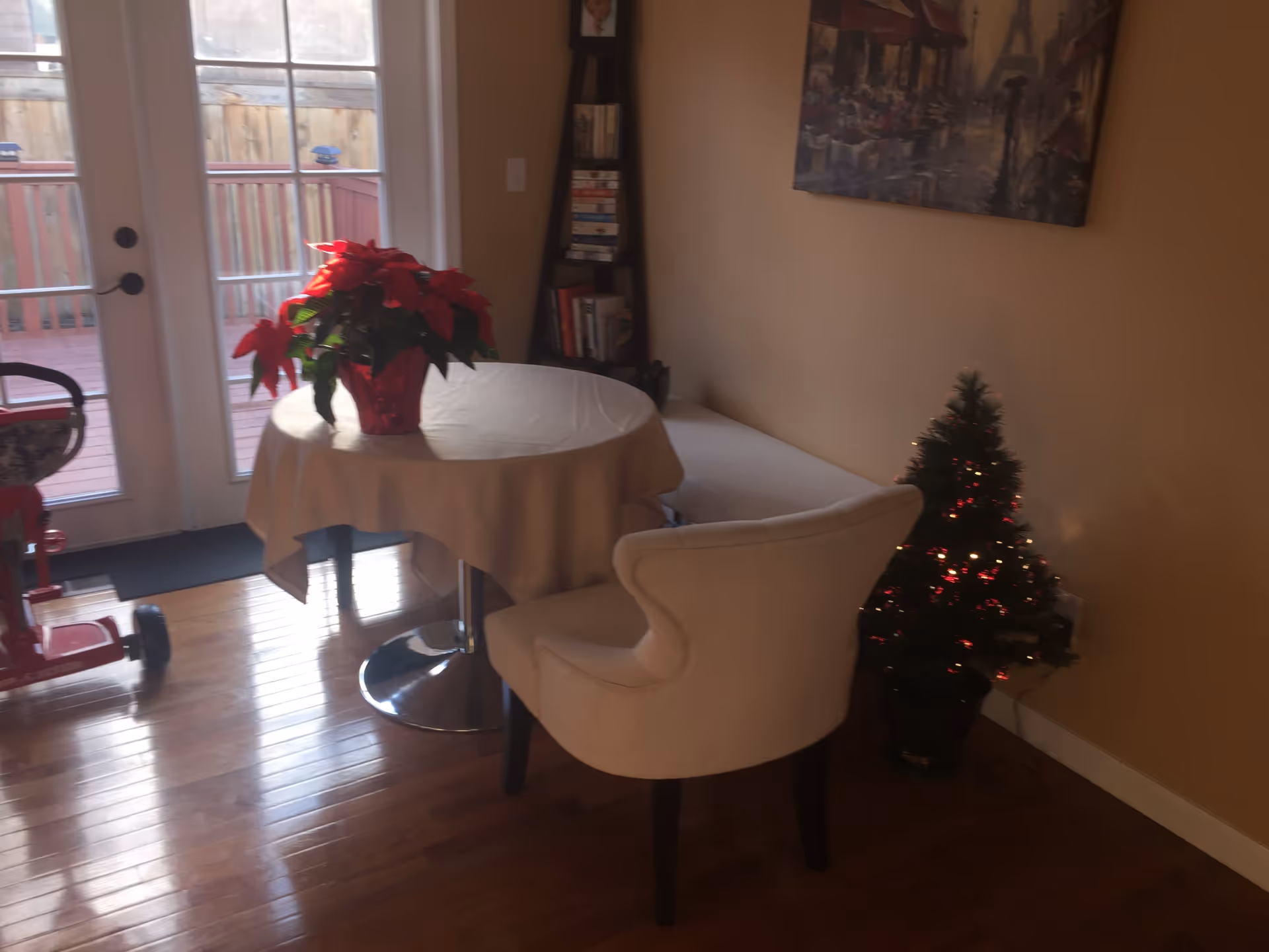 A small dining area with a round table covered by a white tablecloth, a white upholstered chair, a red poinsettia plant on the table, a small decorated Christmas tree on the floor, a bookshelf in the corner, and glass doors leading to an outdoor deck.
