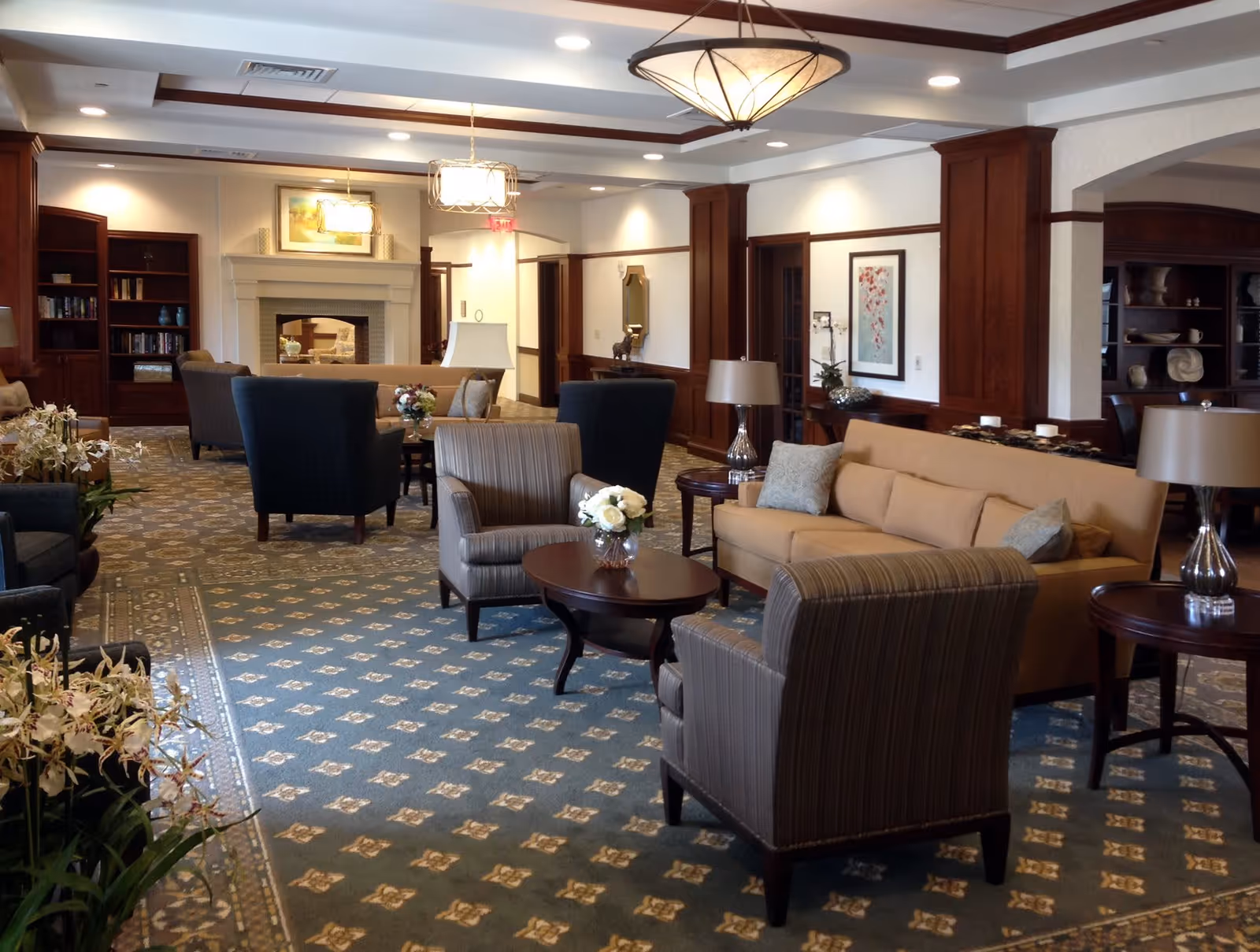 A spacious and well-lit assisted living facility common area with comfortable seating including armchairs and sofas arranged around coffee tables. The room features a patterned carpet, wooden paneling on the walls, built-in bookshelves, decorative lamps, and a fireplace with a framed painting above it. There are also floral arrangements on the tables and plants in the corners, creating a warm and inviting atmosphere.