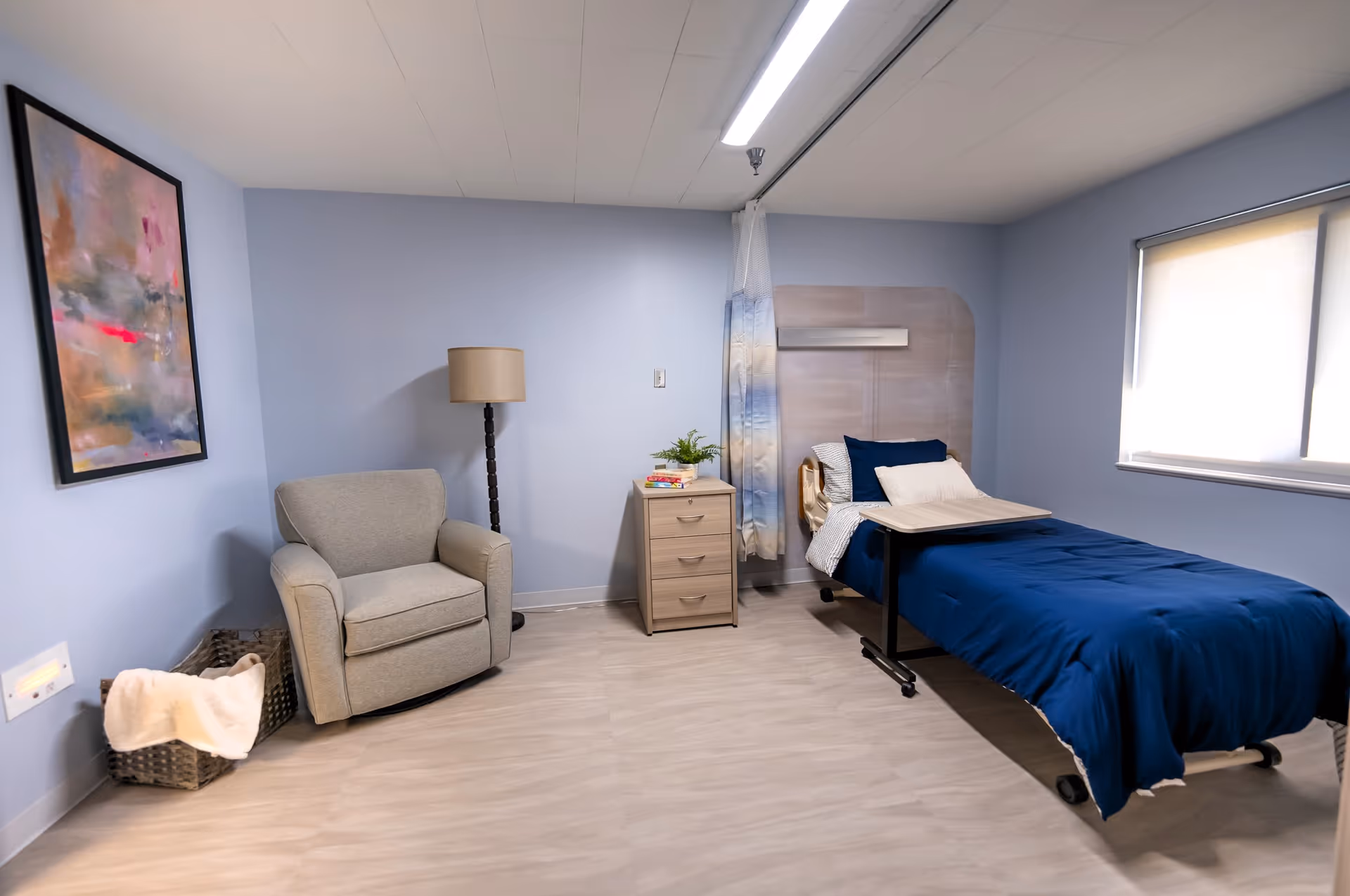A simple, clean bedroom in a senior living facility with light blue walls, a single bed with dark blue bedding, a bedside table with books and a plant, a gray armchair, a floor lamp, a basket with a blanket, and a window with a roller shade.