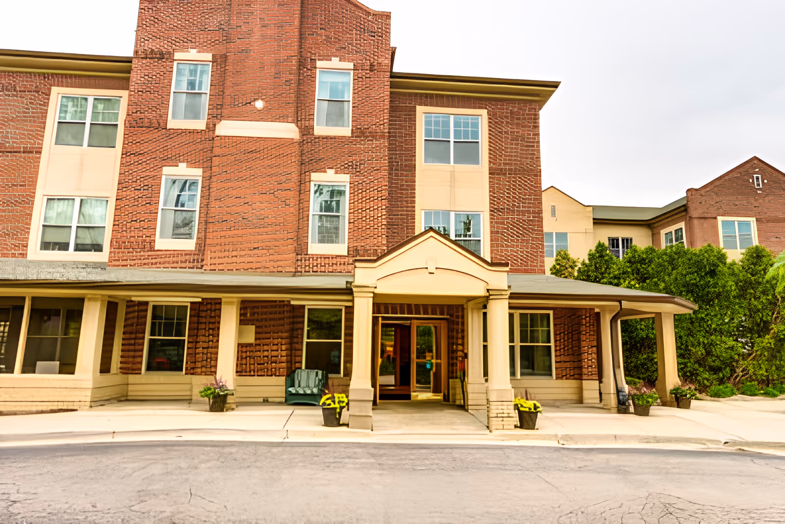 Front entrance of a multi-story brick senior living building with a covered porte-cochere and potted plants.