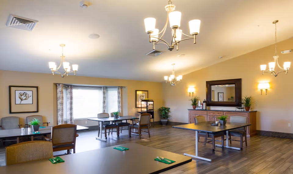 A spacious dining room with several tables and chairs arranged neatly. The room features wooden flooring, beige walls, and multiple chandeliers hanging from the ceiling. There are framed floral artworks on the walls, a large mirror above a wooden sideboard with plants, and large windows with curtains allowing natural light to enter.