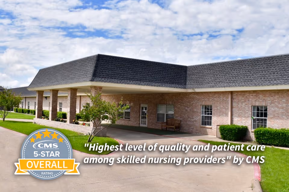 Exterior view of Stonegate Nursing and Rehabilitation facility showing a brick building with a covered entrance, green bushes, small trees, and a driveway under a partly cloudy sky.