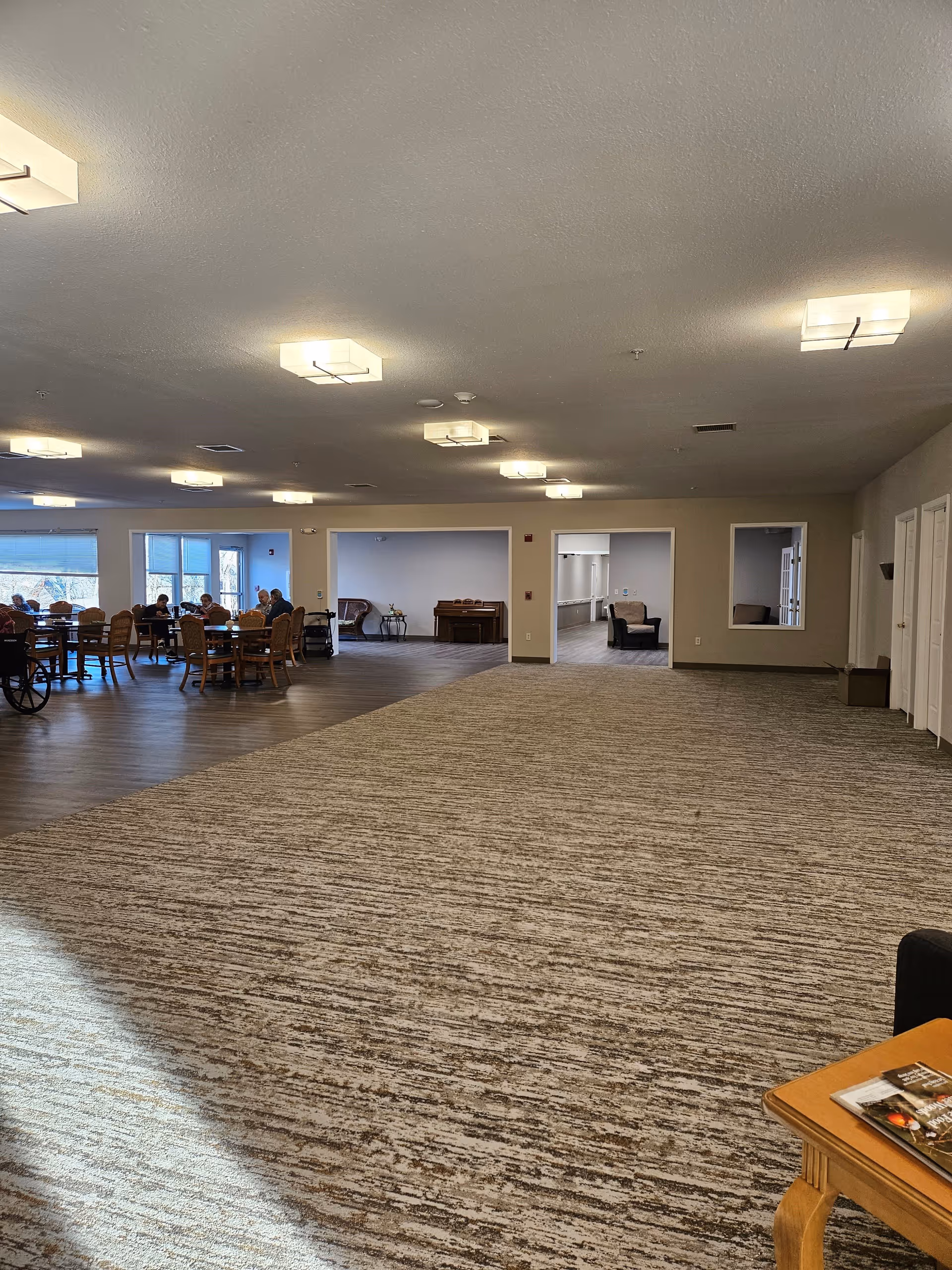 A spacious senior living facility common area with carpeted flooring in the foreground and wood flooring in the background. Several seniors are seated around tables near large windows on the left side. The ceiling has multiple modern square light fixtures. There are three open doorways in the back wall leading to other rooms with chairs and a piano visible. A small wooden table with magazines is in the bottom right corner.