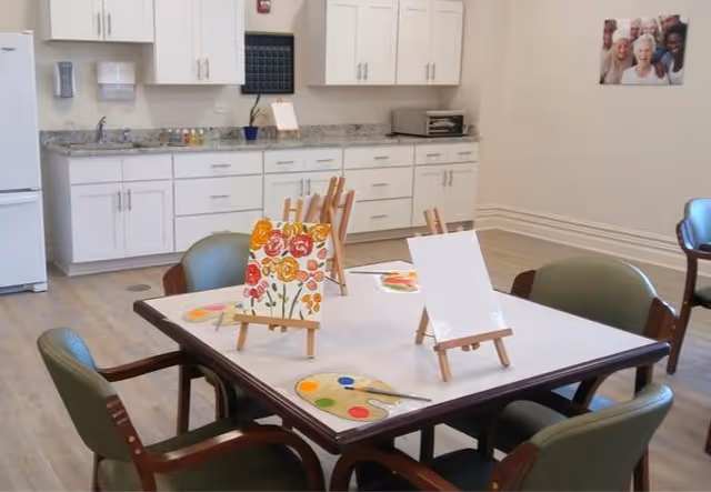 A bright room with a table in the center set up for painting activities, featuring small easels with canvases and paint palettes. The room has white cabinets, a countertop, a refrigerator, and a toaster oven along one wall. There are several chairs around the table and a framed photo of smiling elderly people on the wall.