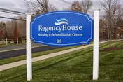 Blue and white outdoor sign for Regency House Nursing & Rehabilitation Center located on a grassy area near a sidewalk and road with trees and power lines in the background.