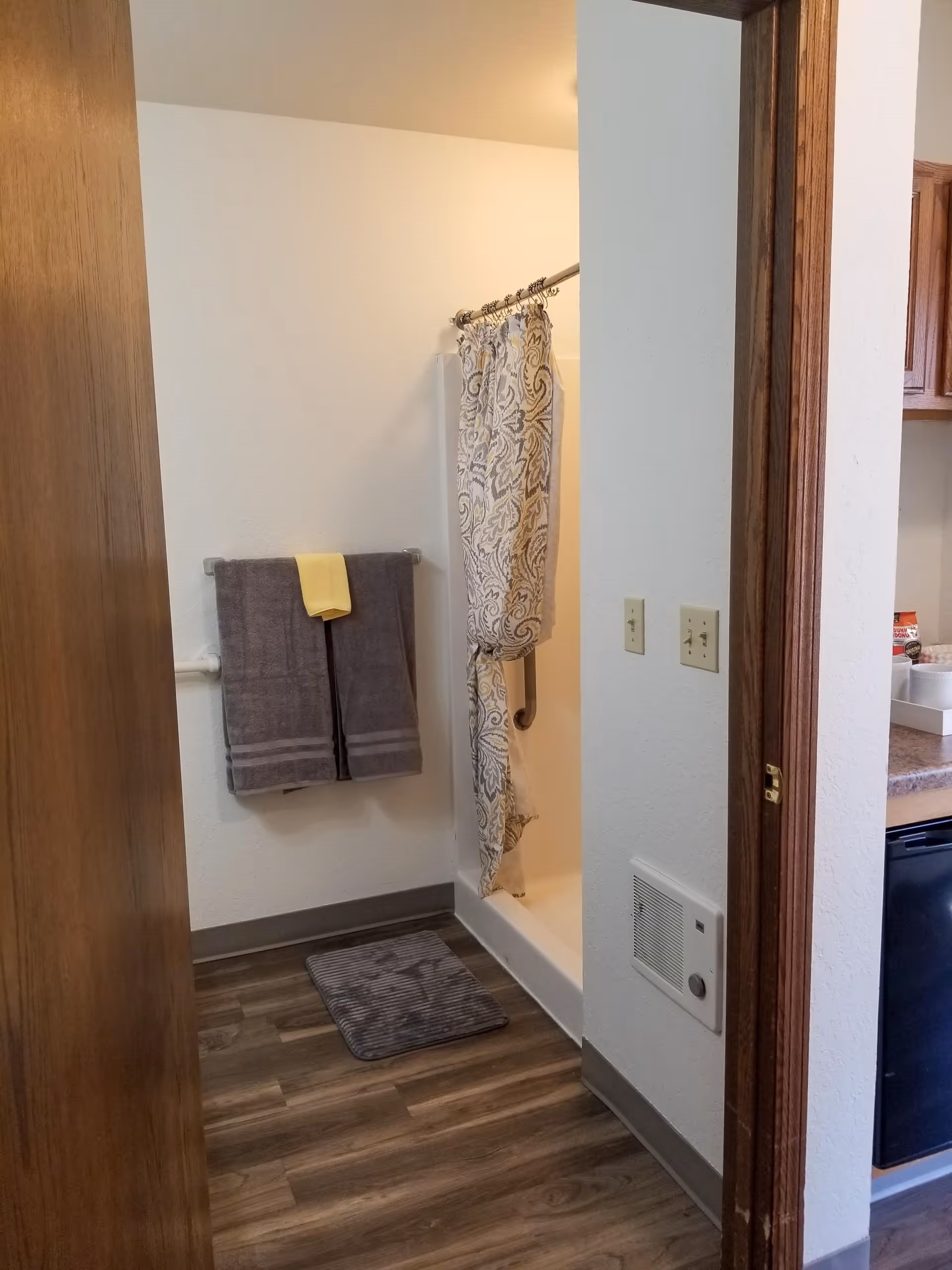 View of a bathroom with a shower area partially covered by a patterned shower curtain. Two gray towels with a yellow towel on top hang on a towel rack on the wall. A gray bath mat is placed on the wooden floor in front of the shower. The bathroom has white walls and a wooden door frame, with a glimpse of a kitchen area visible to the right.