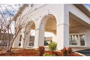 Exterior view of a building entrance with white arches and columns, landscaping with small bushes and a tree without leaves, under a clear sky.