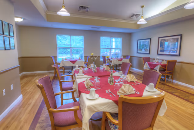 A dining room in a senior living facility with several tables covered in white and red tablecloths, each set with folded napkins, glasses, cups, and silverware. The room has wooden floors, beige walls with a chair rail, framed artwork, and windows letting in natural light.