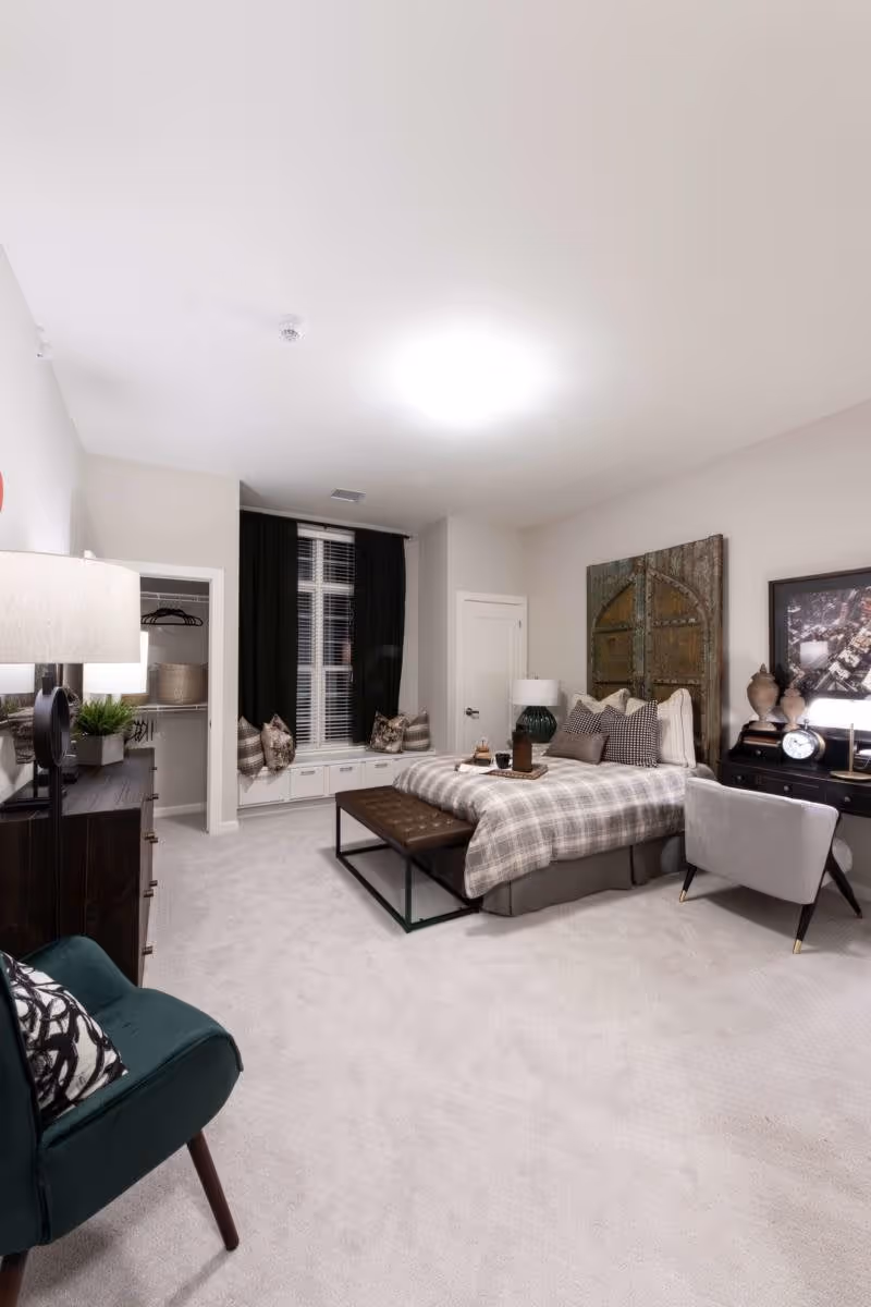 Spacious, well-lit bedroom with a bed dressed in plaid bedding, bench, desk, dresser, window seat and armchairs.