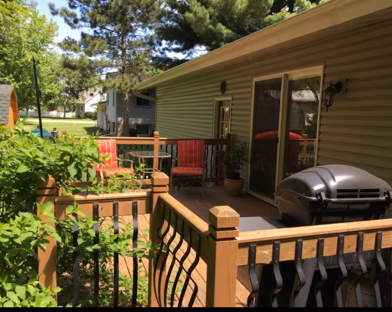 Outdoor wooden deck attached to a house with a grill, two red cushioned chairs, a small round table, potted plants, and a wooden railing with black metal balusters. Green trees and neighboring houses are visible in the background.