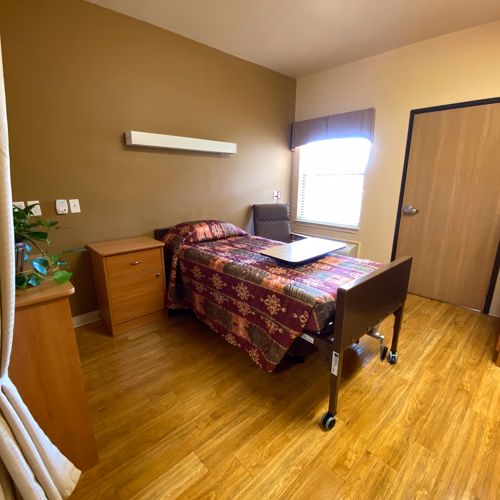 A nursing facility bedroom with a single hospital-style bed covered with a patterned quilt. The room has wooden flooring, a wooden nightstand, a chair near a window with blinds, and a wooden door. There is a plant on a piece of furniture partially visible on the left side.