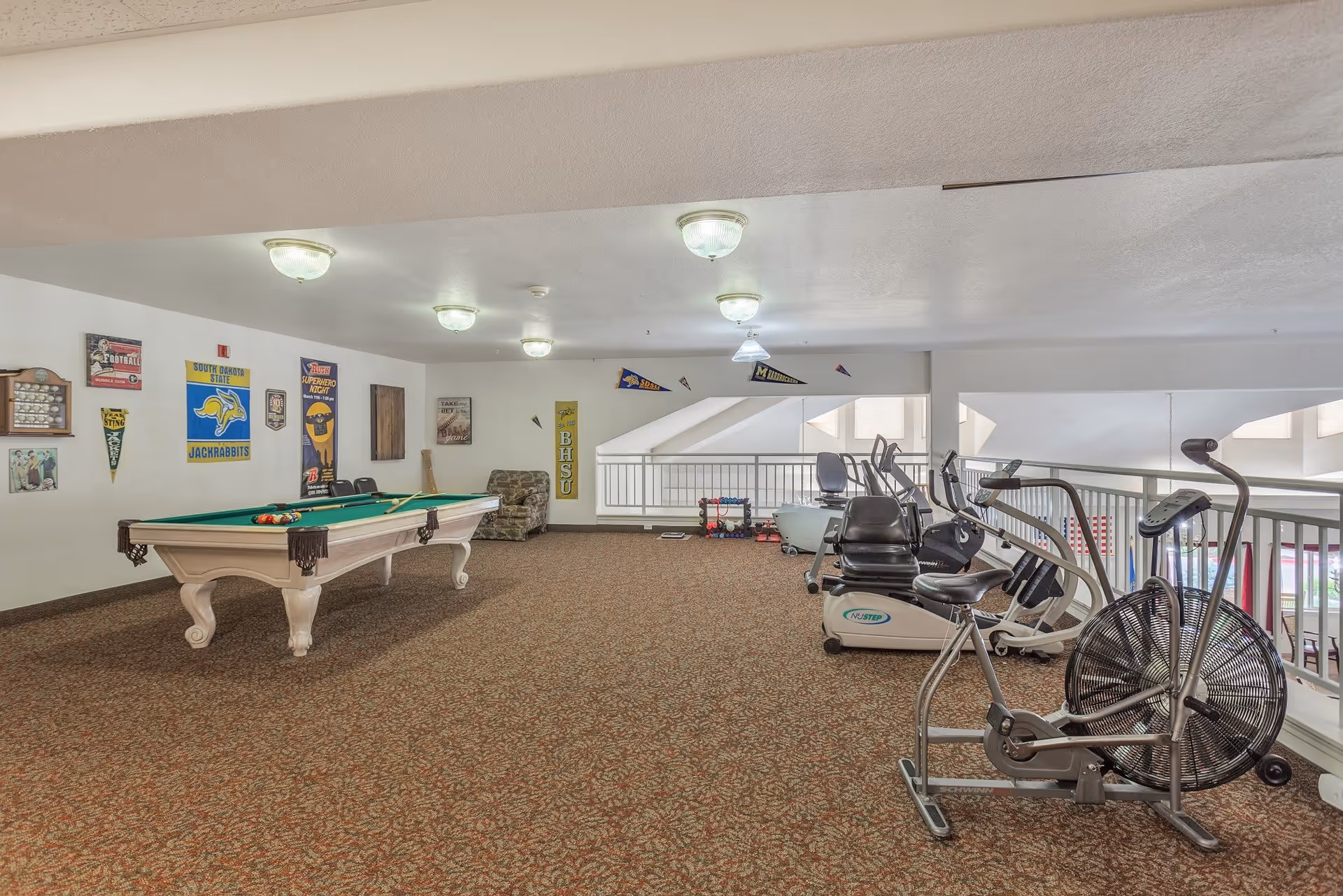 A recreational room with a pool table on the left side and several exercise machines on the right side. The walls are decorated with various sports pennants and posters. The room has carpeted flooring and ceiling lights.