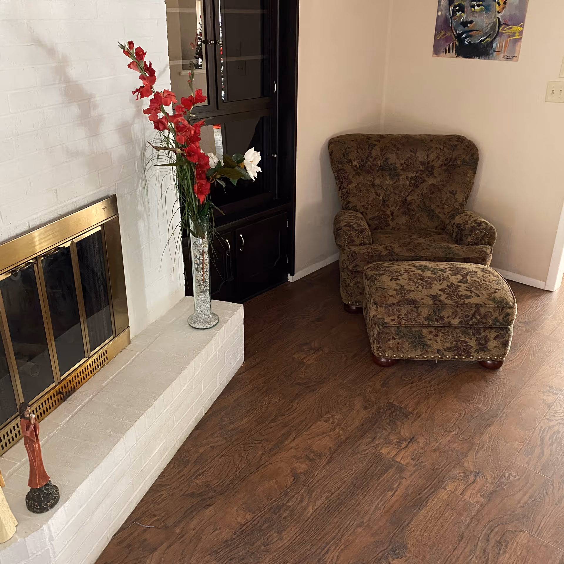 A cozy corner of a living room featuring a floral-patterned armchair with matching ottoman on a wooden floor. To the left is a white brick fireplace with brass-framed glass doors, decorated with a tall vase of red and white flowers and small figurines. A dark wooden cabinet with glass doors is positioned against the wall behind the chair, and a colorful portrait painting hangs on the wall above the chair.