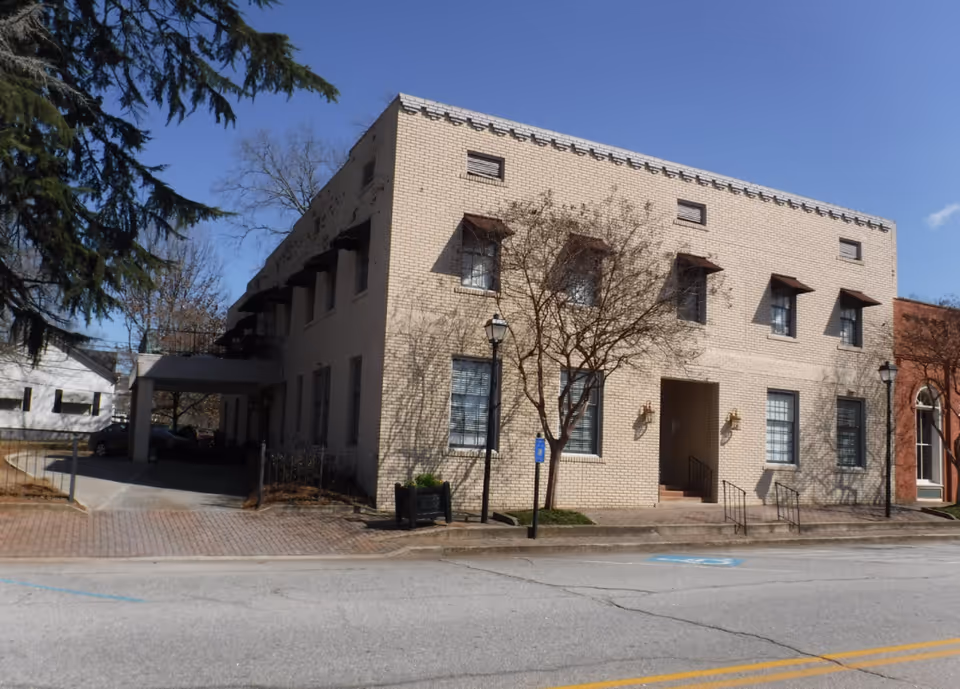 Exterior view of a two-story beige brick building with multiple windows, small awnings above some windows, and a central entrance with stairs. There are leafless trees and street lamps in front, with a clear blue sky overhead.