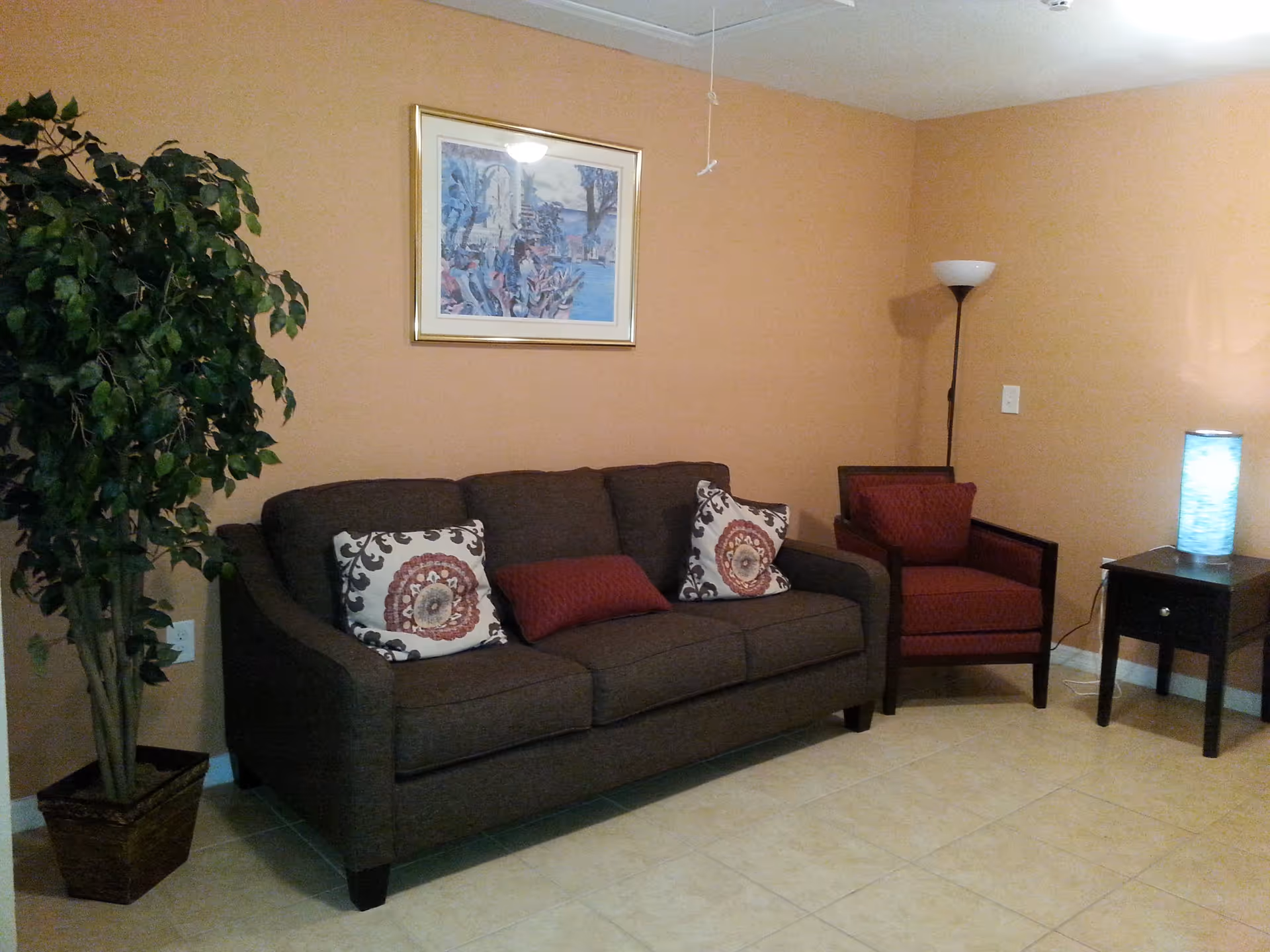 A cozy living room with a brown sofa adorned with three decorative pillows, a red armchair, a tall floor lamp, a small side table with a blue glowing lamp, a large potted plant, and a framed painting on a peach-colored wall.