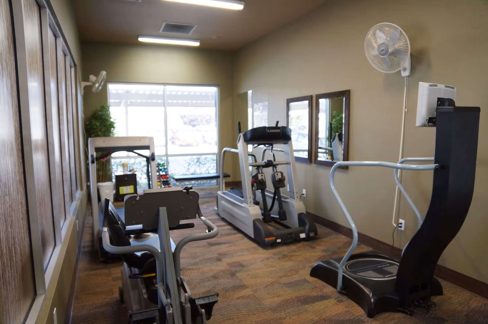 A small indoor exercise room with various fitness equipment including a recumbent bike, a treadmill, and a vibration plate machine. The room has beige walls, a large window letting in natural light, two mirrors on the wall, a wall-mounted fan, and a carpeted floor.