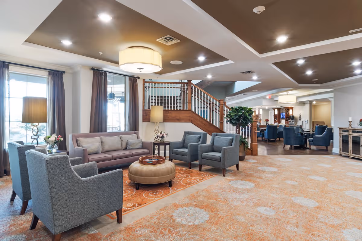 A spacious and well-lit living room area in a senior living facility featuring a seating arrangement with a purple sofa, four gray armchairs, a round beige ottoman, and side tables with lamps and flower vases. The room has large windows with curtains, a patterned carpet, and a staircase with wooden railings leading to an upper floor. In the background, there is a dining area with blue chairs and tables.