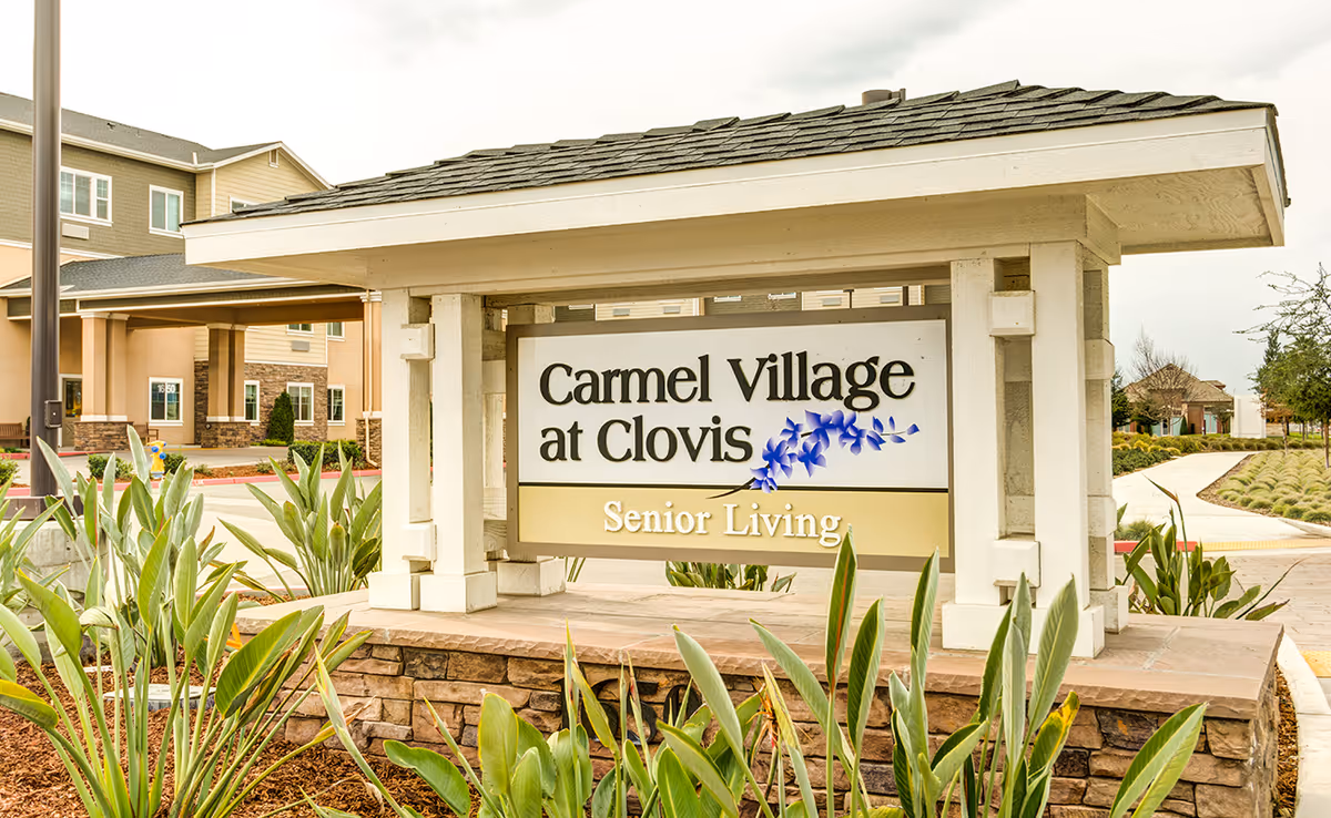 Outdoor entrance sign for Carmel Village at Clovis Senior Living, surrounded by plants and landscaping with part of the building visible in the background under a cloudy sky.