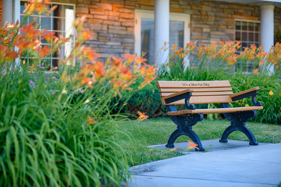 A wooden bench with black metal armrests and legs sits on a concrete path surrounded by green grass and orange flowers. The bench has the inscription 'It's a Marvin Day'. In the background, there is a building with stone walls, white columns, and windows.