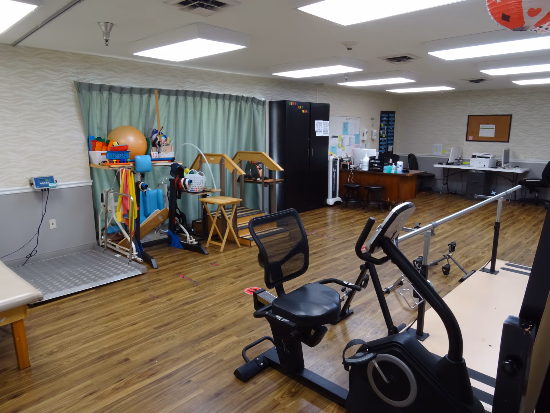 A rehabilitation or physical therapy room with exercise equipment including a stationary bike, parallel bars for walking practice, therapy balls, resistance bands, and other physical therapy tools. The room has wood flooring, fluorescent ceiling lights, a desk with chairs and office supplies, and green curtains covering the windows.