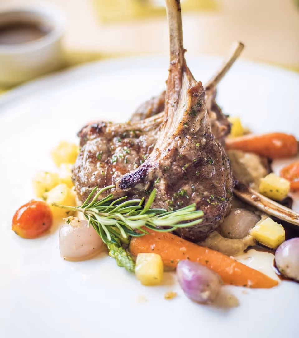Close-up of a gourmet dish featuring two grilled lamb chops garnished with a sprig of rosemary, served with baby carrots, pearl onions, cherry tomatoes, asparagus, and small cubes of yellow vegetables on a white plate.