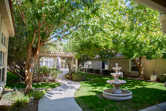A sunny outdoor courtyard with a winding concrete pathway, green grass, leafy trees providing shade, a three-tiered stone fountain, and beige buildings surrounding the area.