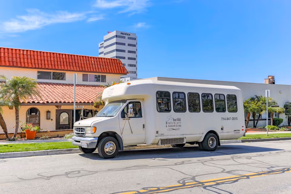 A white Sea Cliff Assisted Living shuttle bus parked on the street in front of a red-tile roof building with palm trees under a clear blue sky.