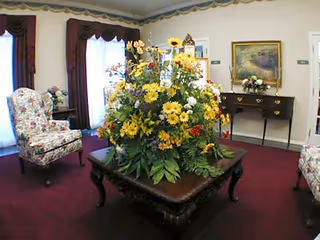 Lobby sitting area with a large floral centerpiece on a coffee table, upholstered chairs, a sideboard, and framed art.