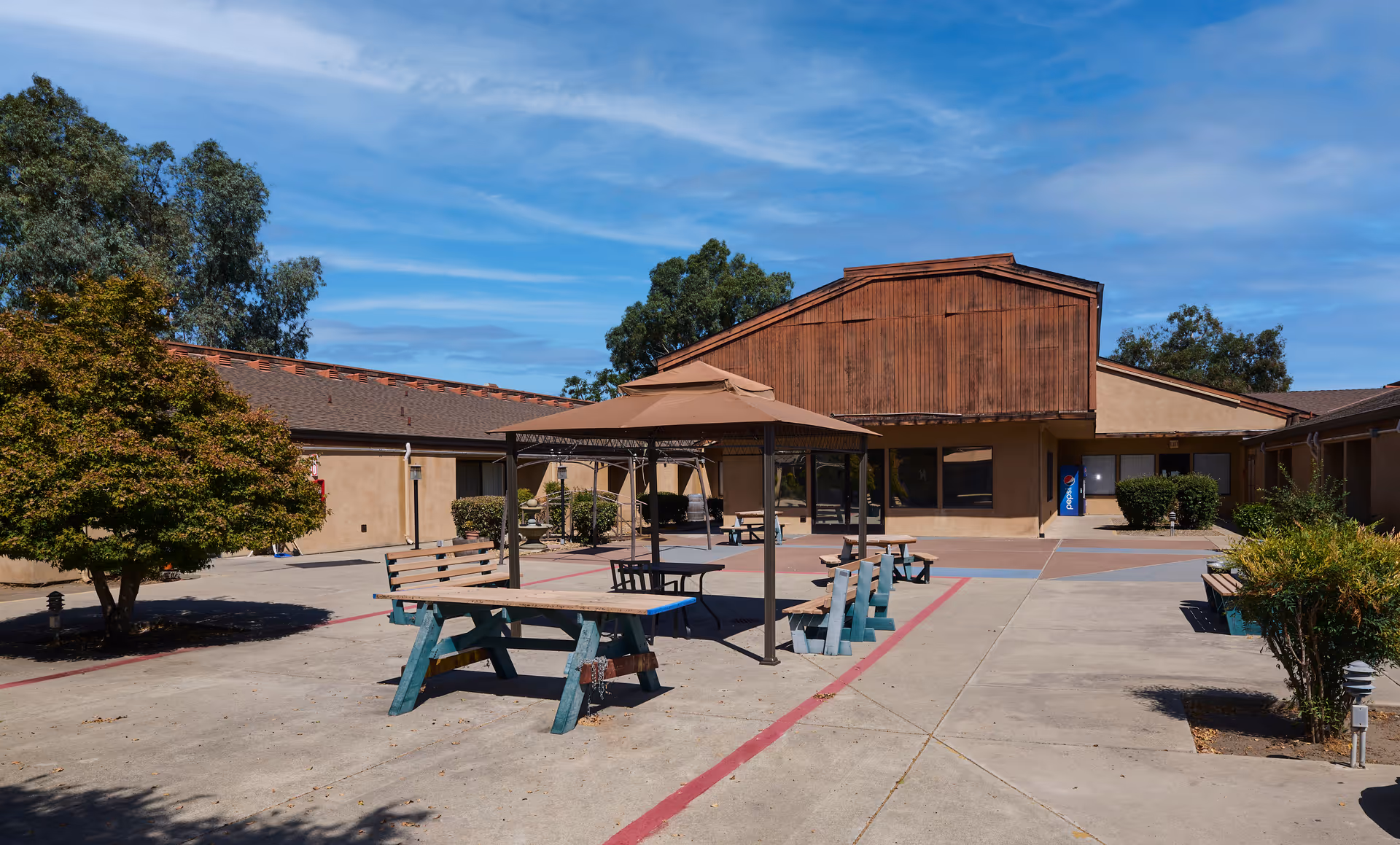 Outdoor courtyard area of Wagner Heights Nursing & Rehabilitation Center featuring picnic tables with benches, a canopy, trees, bushes, and a building with a wooden facade under a partly cloudy blue sky.
