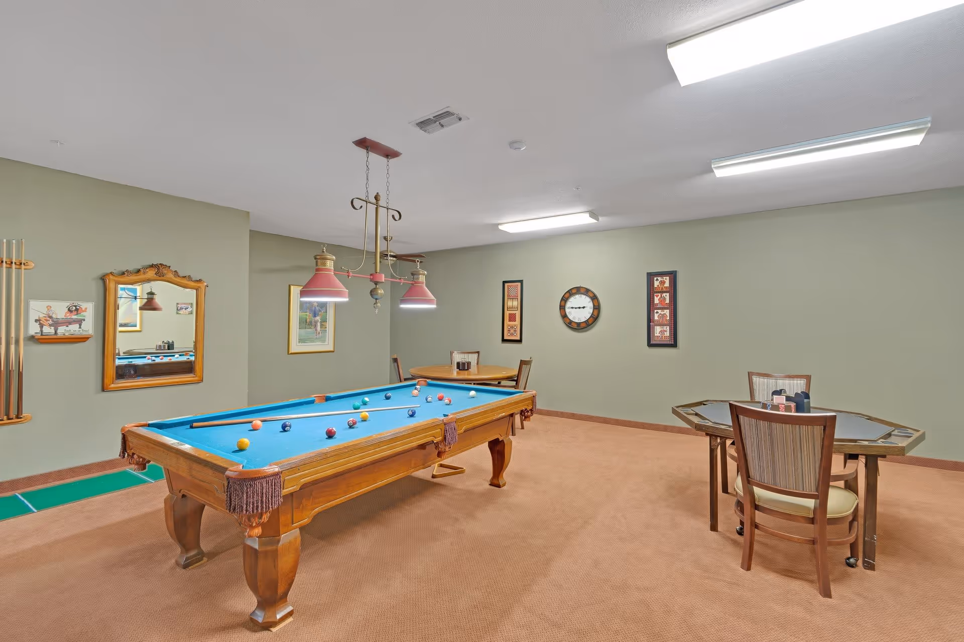 A recreational room with a pool table in the center, a poker table with chairs on the right, and a small round table with chairs in the background. The walls are painted light green and decorated with framed pictures and a clock. The floor is carpeted in a light brown color, and there are overhead fluorescent lights and a hanging light fixture above the pool table.