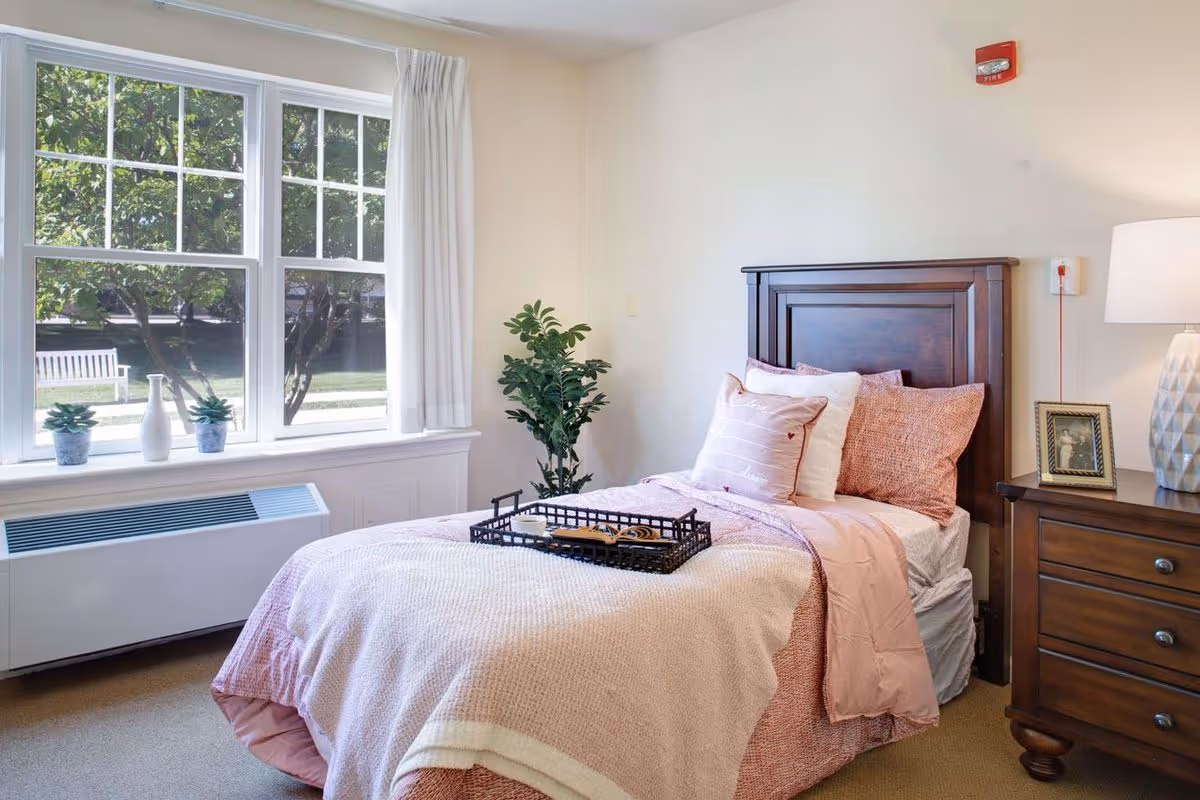 Sunlit bedroom with a single bed dressed in pink linens, a wooden headboard and nightstand next to a large window with potted plants and outdoor greenery visible.