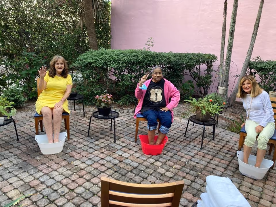 Three women sitting outdoors on chairs with their feet soaking in plastic tubs filled with water. They are smiling and waving, surrounded by plants and a pink wall in the background. Small tables with potted plants are placed between them on a paved patio area.