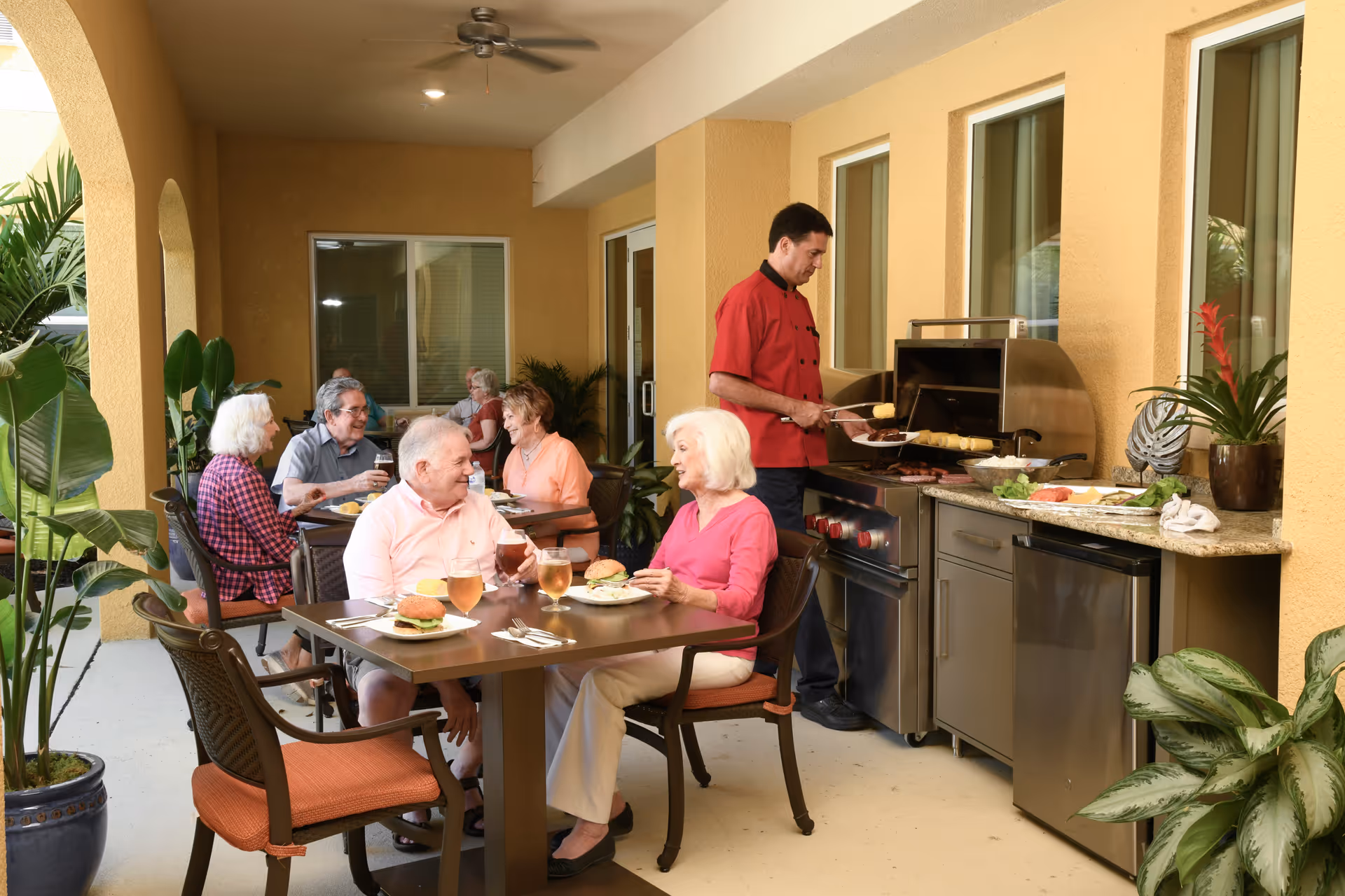 A group of elderly people enjoying a meal and drinks at an outdoor patio dining area. A man in a red chef's jacket is grilling food on a stainless steel barbecue grill. The patio is surrounded by yellow walls and decorated with green potted plants.