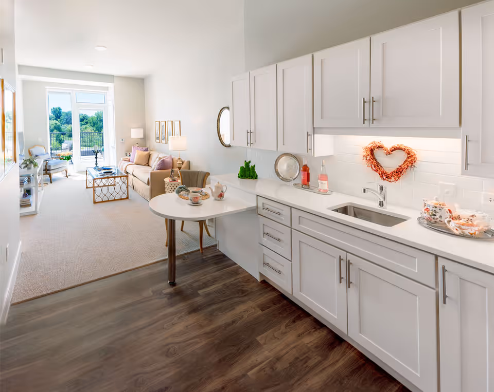 Bright and modern open-concept living space featuring a white kitchen with cabinetry, a small round dining table with chairs, and a cozy living room area with a sofa, armchair, coffee table, and large windows leading to a balcony with greenery outside.