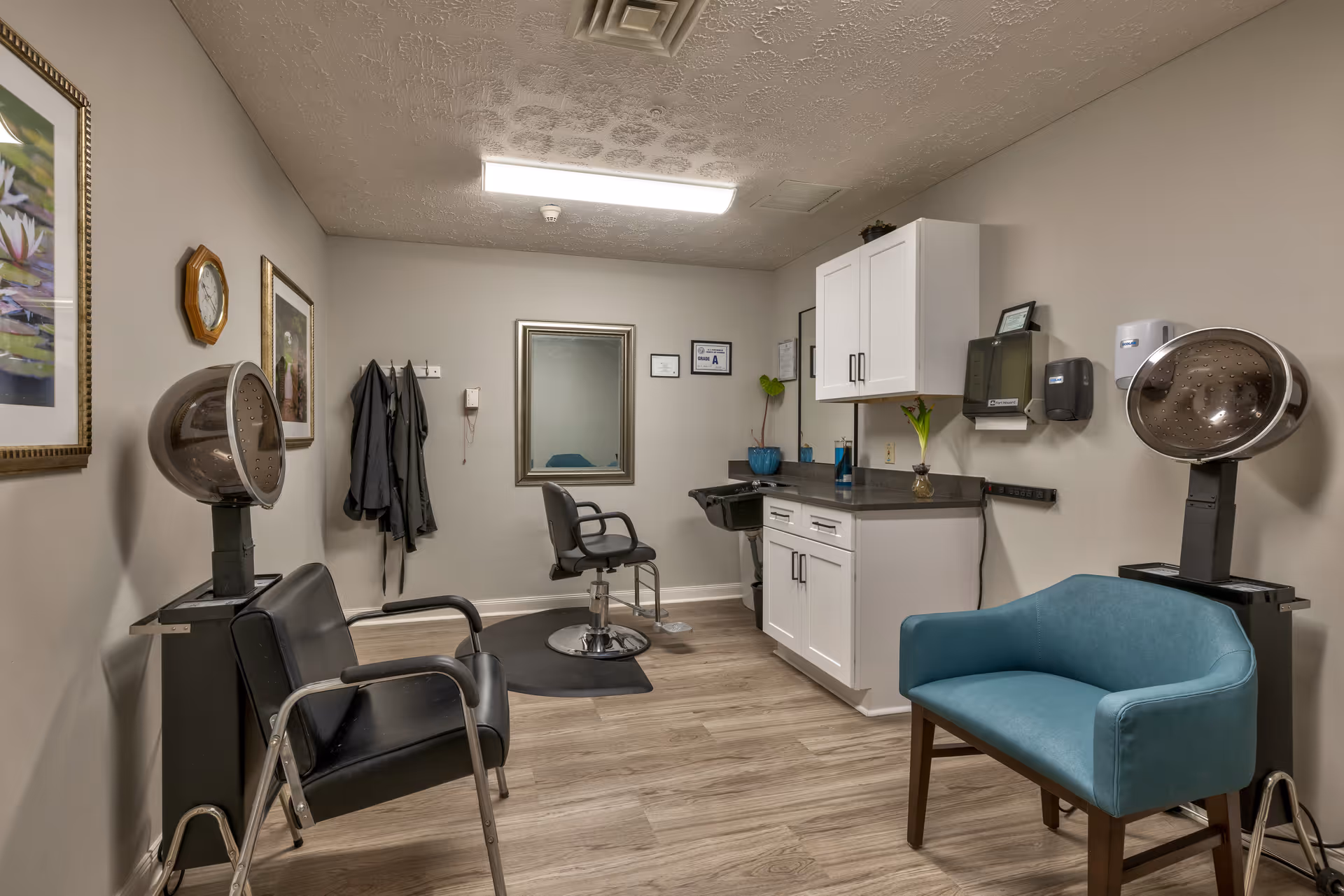 Interior view of a hair salon room in a senior living facility with two black hair drying chairs, a black salon chair in front of a mirror, white cabinets, a sink, a blue cushioned chair, and framed pictures on the walls.