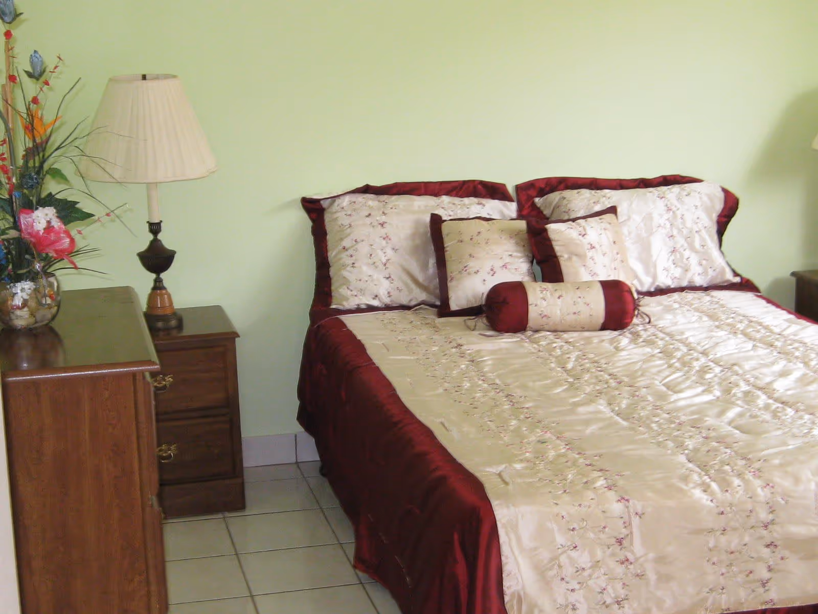 A neatly made bed with cream and burgundy bedding beside a wooden nightstand, lamp, and dresser topped with a floral arrangement.
