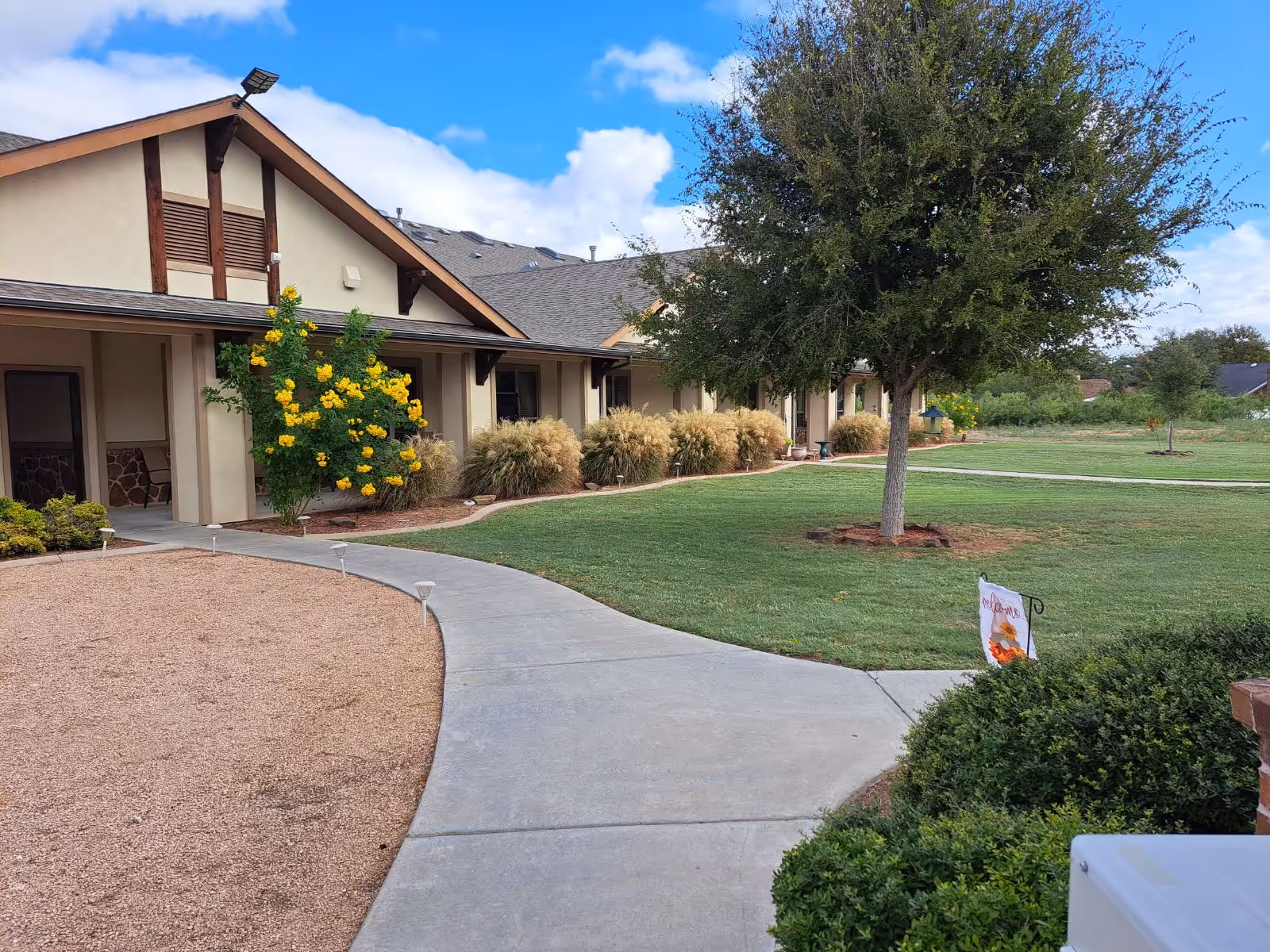 Exterior view of a single-story senior living building with a curved concrete walkway, green lawn, trees, and flowering shrubs.