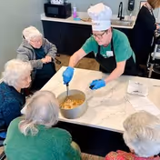 A staff member wearing a chef hat and gloves is serving food from a pot on a kitchen island to a group of elderly women seated around the island in a senior living facility kitchen area.