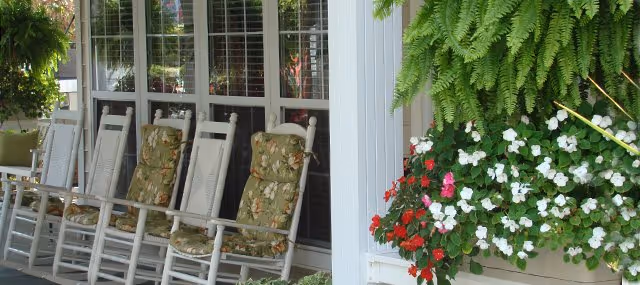 A row of white wooden rocking chairs with floral cushions lined up on a porch in front of large windows. To the right, there is a hanging fern and a planter box filled with red, white, and pink flowers. The setting appears calm and inviting.
